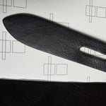 Load image into Gallery viewer, A close-up of a black textured leather strap with an oval cutout, laid diagonally on a white textured surface with faint geometric line drawings.