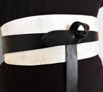 Load image into Gallery viewer, A close-up of a wide white belt with a black leather strap and a black leather bow, worn over a black ribbed top. The white belt features a minimalist repeating geometric line pattern.