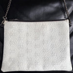Load image into Gallery viewer, A white textured leather clutch bag with a subtle geometric monogram pattern, featuring a silver-tone chain strap and a top zipper, displayed on a black leather background.

